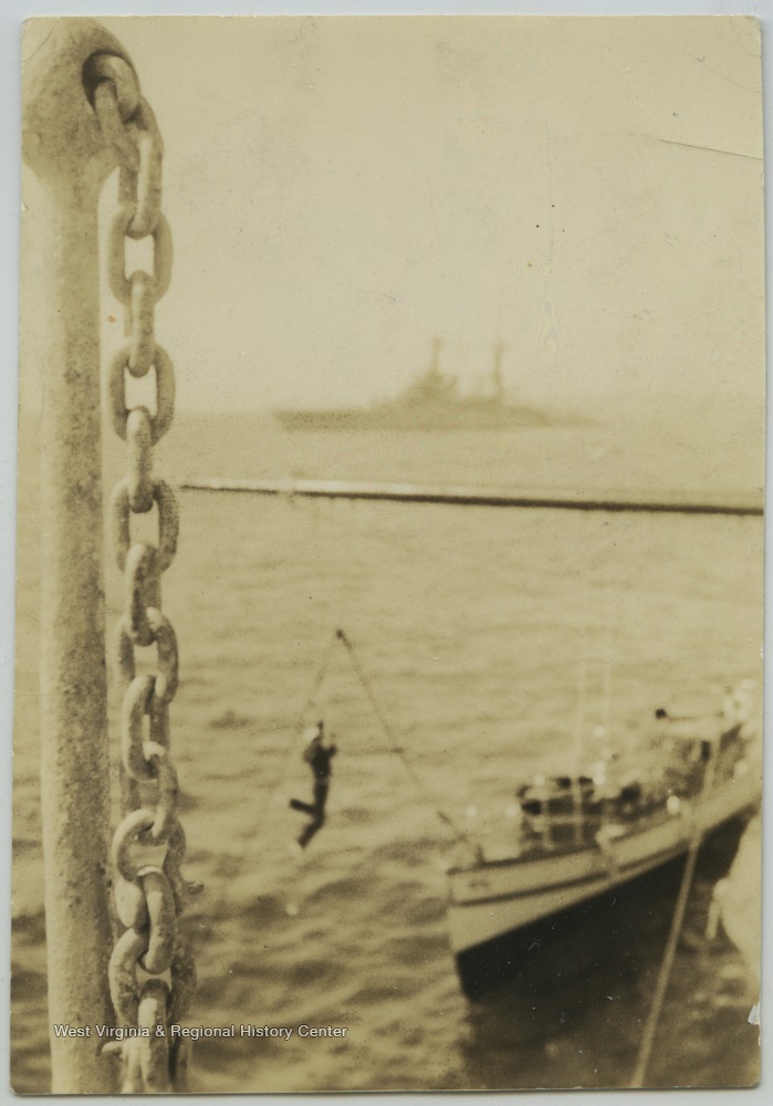 A sailor is transferred to the motor boat by gliding down a rope and pulley system. Motor boats were used to carry enlisted men ashore. Photo taken from the deck of the U.S.S. West Virginia. An unidentified battleship lurks in the background. 
